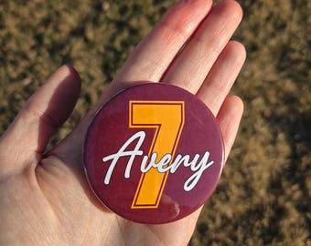 Könnte beinhalten: Ein runder Button mit weinrotem Hintergrund, der den Namen "Avery" in weißer Schrift und die Zahl "7" in Gold zeigt. Der Button wird in einer Hand gehalten, vor einem verschwommenen Hintergrund im Freien.