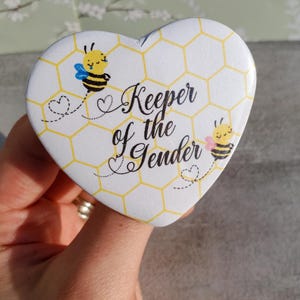 May include: Heart-shaped button with a white background and a honeycomb pattern. The button features two cartoon bees and the text "Keeper of the Gender" in black script. The bees have yellow bodies, black stripes, and blue or pink wings.
