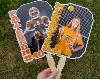 Könnte beinhalten: Zwei fächerförmige Handfächer mit Sportmotiven. Einer zeigt einen Footballspieler in orangefarbener Uniform mit dem Namen "PATTERSON #40". Der andere zeigt eine Softballspielerin in einem orangefarbenen Trikot mit dem Namen "MCKENZIE #14". Beide Fächer haben Holzgriffe.