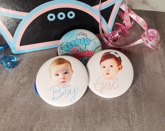 May include: A collection of baby shower decorations, including two round buttons with baby faces and the text "Team Boy" and "Team Girl." Other items include a pacifier, a banner, and a button with the text "Keeper of the Under." The color scheme is blue, pink, and white.