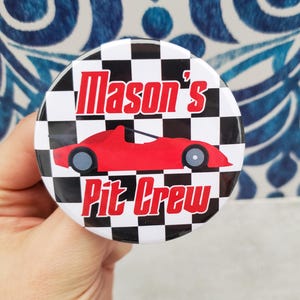 May include: A round button with a black and white checkered background. The button features the text "Mason's Pit Crew" in red and a red race car graphic. The button is held in a hand.