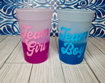 May include: Two plastic cups with a color gradient. The cup on the left is pink and says "Team Girl" in pink lettering. The cup on the right is blue and says "Team Boy" in blue lettering.