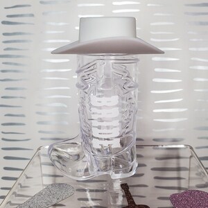 K&ouml;nnte beinhalten: Ein durchsichtiges Cowboy-Stiefel-Schnapsglas aus Plastik mit einem wei&szlig;en Cowboyhut oben drauf. Der Stiefel hat das Wort "Howdy" an der Seite. Das Schnapsglas steht auf einer durchsichtigen Glasfl&auml;che mit einer braunen Gitarre und einem lila glitzernden Cowboyhut.