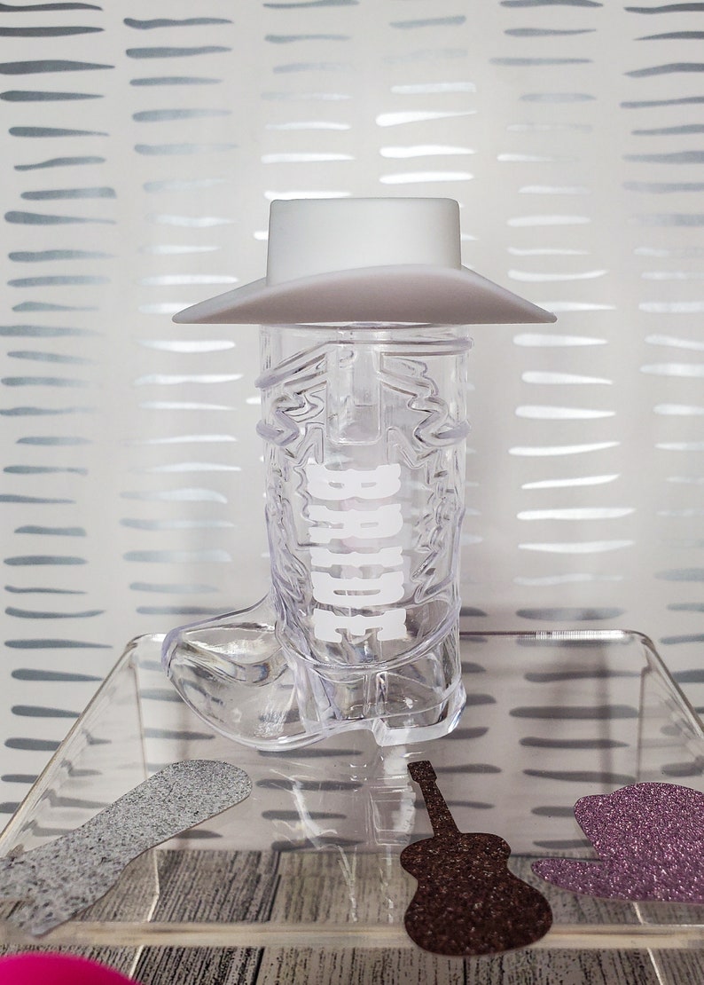 K&ouml;nnte beinhalten: Ein durchsichtiges Cowboy-Stiefel-Schnapsglas aus Plastik mit einem wei&szlig;en Cowboyhut oben drauf. Der Stiefel hat das Wort "Howdy" an der Seite. Das Schnapsglas steht auf einer durchsichtigen Glasfl&auml;che mit einer braunen Gitarre und einem lila glitzernden Cowboyhut.