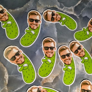 May include: A collection of green pickle-shaped stickers, each featuring a smiling face wearing sunglasses. The stickers have a white border and are scattered across a marbled surface. The pickle design includes small, dark green details.