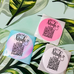 May include: Three round stickers with the text "BUY THE bride A DRINK" and a ring emoji. Each sticker has a different background color: pink, blue, and white. The stickers have a QR code.