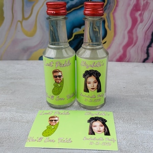 May include: Two small glass bottles with red caps and custom labels. One label says "Last Pickle" with a cartoon pickle and a man's face. The other says "Elizabeth's" with a woman's face. A matching card is below.