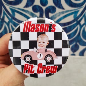 May include: A round button with a black and white checkered background. The button features the text "Mason's Pit Crew" in red and a cartoon image of a baby in a pink race car. The car has the number 1 on it.