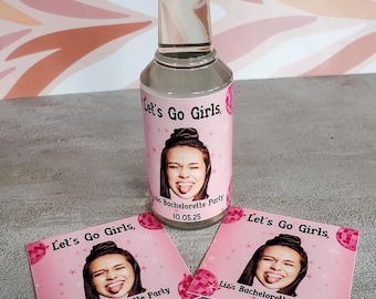 May include: A clear glass bottle with a red cap and a pink label that reads "Let's Go Girls." The label features a photo of a woman sticking out her tongue. Two matching pink coasters are also present.