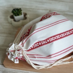 Może przedstawiać: Biała torba z tkaniny z czerwonymi haftowanymi paskami i białym sznurkiem do ściągania. Torba leży na drewnianej powierzchni.