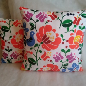 以下が含まれることがあります： 白い背景にカラフルな花柄が描かれた、装飾的なクッションが2つ。柄には、赤、オレンジ、黄色、緑、青、紫の花が含まれています。クッションの1つには、小さな青い鳥が中央に描かれています。