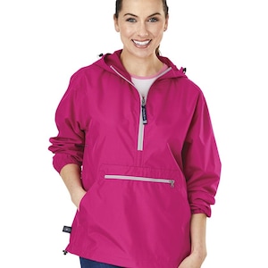 May include: A bright pink anorak jacket with a hood and a half-zip closure. The jacket has a front pocket with a silver zipper and a small logo tag on the side. The model is wearing blue jeans.