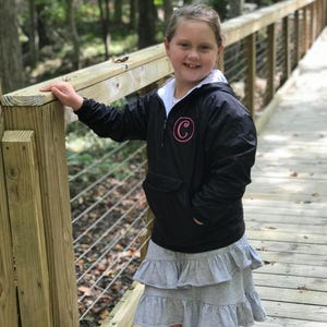 May include: A young person wearing a black hooded jacket with a pink letter "C" on the chest, a white undershirt, and a gray tiered skirt. The person is standing on a wooden bridge in a forest setting.