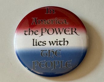 May include: A round button with a red, white, and blue gradient background. The text "In America, the POWER lies with THE PEOPLE" is printed in black, stylized font. The button is a patriotic item.