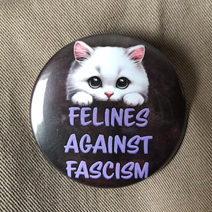 May include: A white cat with pink ears peeks over the top of a black button with the text "FELINES AGAINST FASCISM" in purple letters.