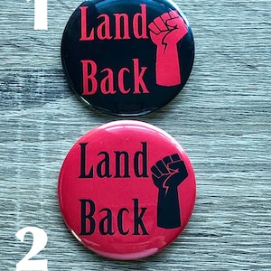 Op de afbeelding: Twee ronde knoppen met de tekst "Land Back" en een gebalde vuist. De eerste knop is zwart met rode tekst en een rode vuist. De tweede knop is rood met zwarte tekst en een zwarte vuist.