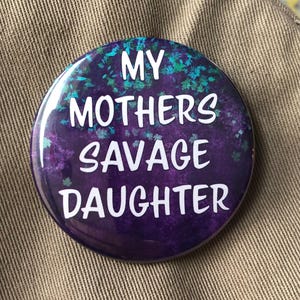 May include: A purple button with a white background and the text "My Mothers Savage Daughter" in white lettering.