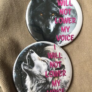 May include: Two round buttons with a wolf design. The top button features a black wolf with yellow eyes and the text "I WILL NOT LOWER MY VOICE" in pink. The bottom button features a white wolf howling at the moon with the same text in pink.