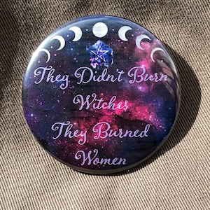 May include: A round button with a galaxy background and a pentagram in the center. The text on the button reads "They Didn't Burn Witches They Burned Women".