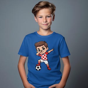 Boy wearing blue t-shirt with cartoon soccer player dabbing, standing by a soccer ball.