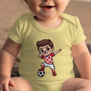 Baby wearing a yellow onesie with a cartoon boy dribbling a soccer ball.