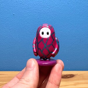 May include: A magenta and teal figurine of a character from the video game Fall Guys. The figure has a rounded body, small arms, and a white face with two black eyes. It is standing on a purple base.