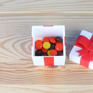 May include: A white gift box with a red ribbon and bow. The box is open and filled with a variety of colorful candies.