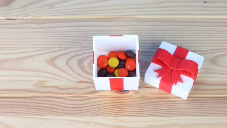 May include: A white gift box with a red ribbon and bow. The box is open and filled with a variety of colorful candies.