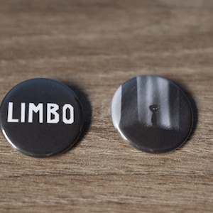 May include: Two black and white button pins. One pin has the word "LIMBO" in white text. The other pin has a black and white image of a figure standing in a forest.