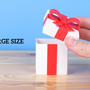 May include: A white gift box with a red ribbon and bow is being opened by a hand. The box is sitting on a wooden surface. The text "LARGE SIZE" is visible in the background.