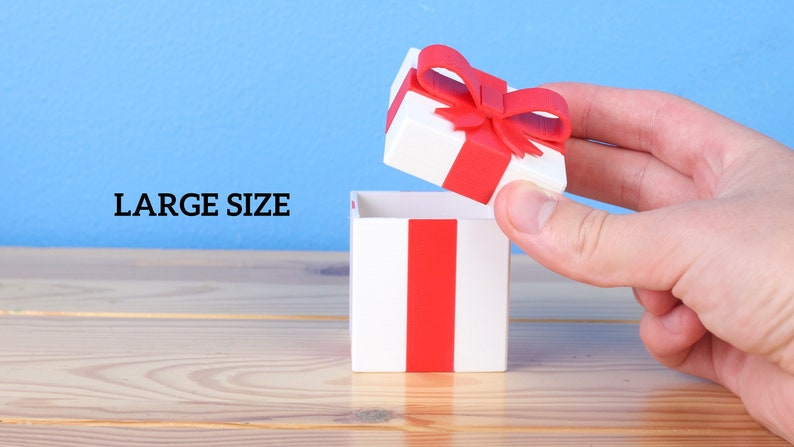 May include: A white gift box with a red ribbon and bow is being opened by a hand. The box is sitting on a wooden surface. The text "LARGE SIZE" is visible in the background.