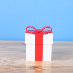 May include: A white gift box with a red ribbon and bow. The box is sitting on a wooden surface.