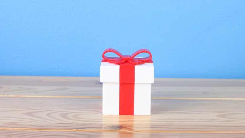 May include: A white gift box with a red ribbon and bow. The box is sitting on a wooden surface.