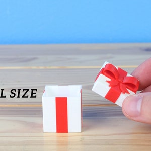 May include: A small, white gift box with a red ribbon and bow. The box is open and the lid is being held by a hand. The text "SMALL SIZE" is visible in the image.