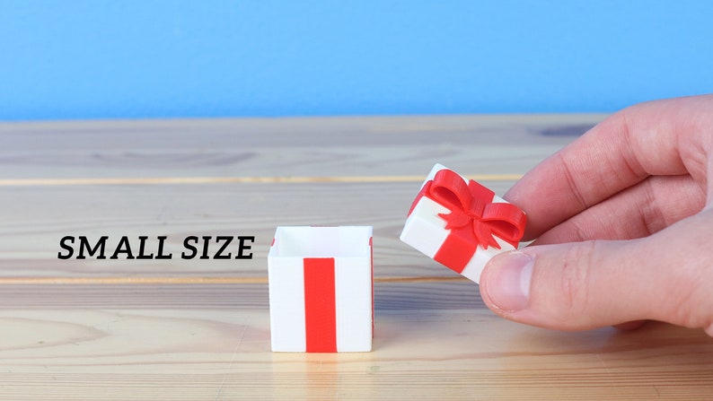 May include: A small, white gift box with a red ribbon and bow. The box is open and the lid is being held by a hand. The text "SMALL SIZE" is visible in the image.