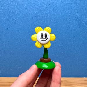 May include: A yellow flower figurine with a smiling face, green stem, and a green and brown base. The flower's petals are bright yellow, and the face has a black outline with a white center. The figurine is a collectible item.
