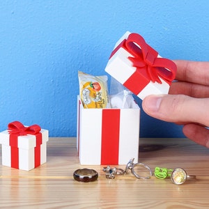 May include: A white gift box with a red ribbon and bow. The box is open and contains a small package of fruit snacks. There are also several rings on the table next to the box.