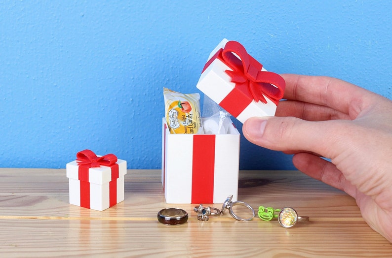May include: A white gift box with a red ribbon and bow. The box is open and contains a small package of fruit snacks. There are also several rings on the table next to the box.