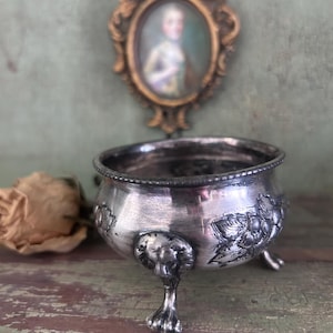 May include: An antique silver bowl with three decorative lion's paw feet. The bowl has embossed floral patterns and a beaded rim. A vintage portrait in a gold frame is in the background, with a dried rose on the left.