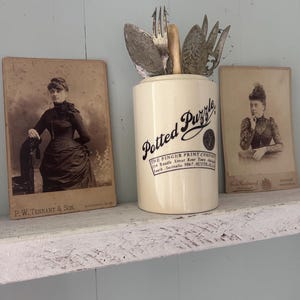 May include: A cream ceramic jar with the text "Potted Puzzle" and vintage silverware. Two antique photographs sit either side of the jar on a distressed white shelf against a light blue wall. The jar has a black logo and text.
