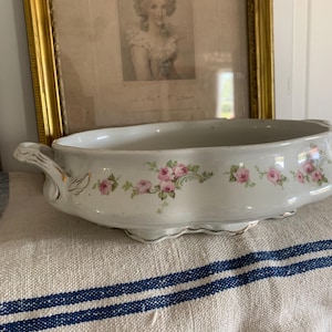 May include: White porcelain serving dish with pink rose floral pattern and gold trim. The oval dish has handles and sits on small feet. A framed portrait hangs in the background.