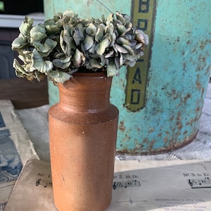 May include: A small, brown, ceramic vase with a dried, grey hydrangea bouquet. The vase is sitting on a sheet of music with the words "How Lovely Are Thy Dwell" printed on it.