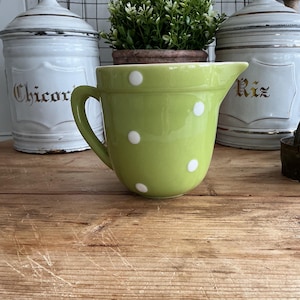 May include: A lime green ceramic jug featuring a pattern of white polka dots. The jug has a curved handle on the left side and a pouring spout on the right side. The surface of the jug is smooth and glossy, reflecting light.