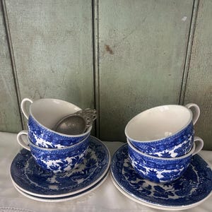 May include: Four blue and white porcelain teacups and saucers with a Willow pattern design. A silver tea infuser rests in one of the teacups. The teacups and saucers are stacked on a white surface.