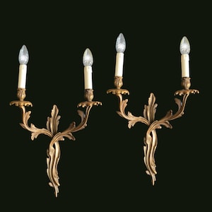 May include: A pair of gold-toned metal wall sconces, each featuring two candle-style light fixtures with white sleeves. The sconces have an ornate, scrolling leaf design that extends from a central point, supporting two candle cups on curved arms.