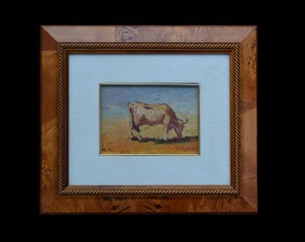 May include: A framed oil painting of a brown and white cow grazing in a field. The painting is set against a blue sky with white clouds.