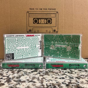 May include: Cassette tapes with green maze designs, labeled "Cassette Labyrinth: The Shining Edition." The tapes have clear cases and feature the text "Overlook Hedge Maze." A cardboard box with a cassette tape illustration and the text "Thank you for your purchase" is in the background.