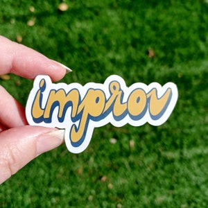 May include: A white sticker with the word "improv" in a retro font. The text is yellow with a dark blue outline, set against a white background. The sticker is held against a backdrop of green grass.