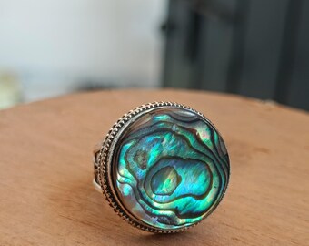 Könnte beinhalten: Ein silberner Ring mit einem runden Einlegeteil aus Abalone-Muschel. Die Muschel zeigt ein wirbelndes Muster in Grün, Blau und schillernden Farben.