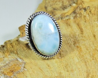 Op de afbeelding: Een zilveren ring met een grote, ovale, lichtblauwe edelsteen. De edelsteen is gezet in een gedraaide, touwachtige rand. De band van de ring is van effen zilver. De ring wordt getoond op een lichtbruin oppervlak.